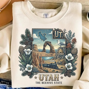 Könnte beinhalten: Cremefarbenes Sweatshirt mit einer detaillierten Grafik der Landschaft von Utah, einschließlich Delicate Arch, einem Zug und einer Möwe. Das Design wird von floralen und Kiefernelementen umrahmt, mit dem Text "UTAH" und "THE BEEHIVE STATE".