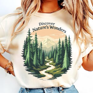 Pode incluir: Camiseta creme com um gráfico temático da natureza. O design apresenta um rio sinuoso através de uma floresta de árvores verdes, com montanhas ao fundo. O texto "Discover Nature's Wonders" está arqueado acima da cena.