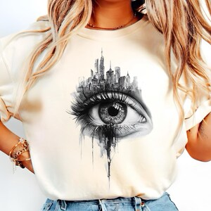 Puede incluir: Una camiseta blanca con un estampado gráfico en blanco y negro de un horizonte de ciudad dentro de un ojo. El ojo está dibujado en un estilo realista con pestañas y una lágrima.