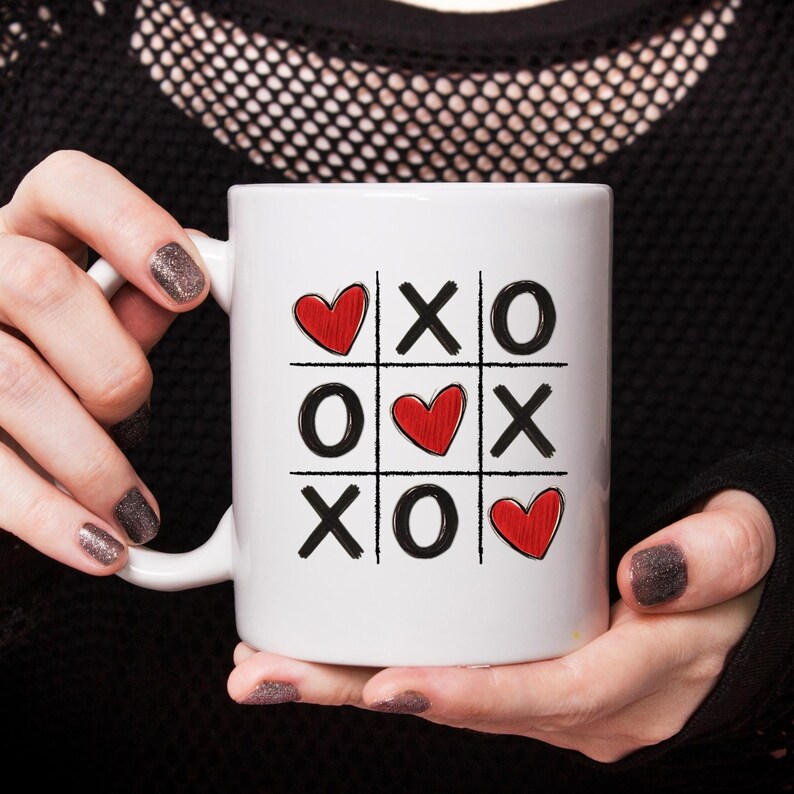 May include: White ceramic mug with a black and red tic-tac-toe design. The design features three red hearts and the letters X and O.