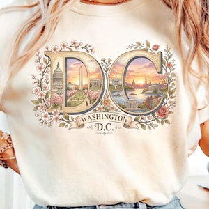 Pode incluir: Camiseta creme com um gráfico colorido do horizonte de Washington D.C. dentro das letras "DC". O design inclui o Capitólio, o Monumento a Washington e uma cena de pôr do sol, rodeado por elementos florais e o texto "WASHINGTON D.C."