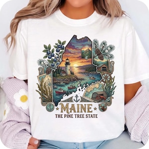 May include: White t-shirt featuring a colorful graphic of the state of Maine. The design includes a lighthouse, boats, maps, and the text "MAINE THE PINE TREE STATE". The graphic is surrounded by illustrations of blueberries, pine cones, and foliage.