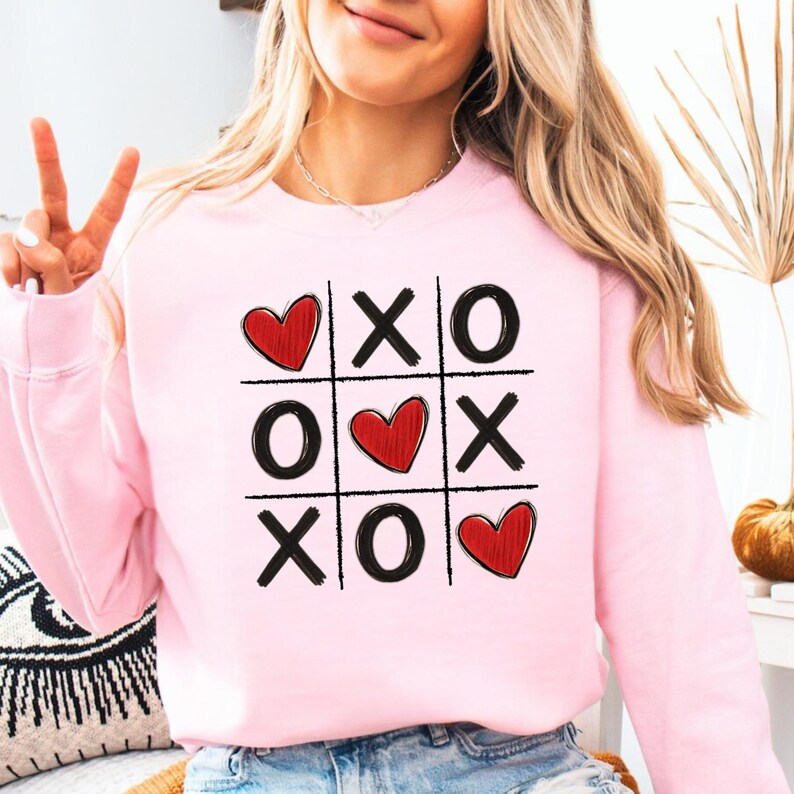 May include: Pink sweatshirt with a graphic of a tic-tac-toe board with hearts and X's and O's.