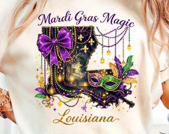 May include: Cream-colored t-shirt featuring a Mardi Gras design. The design includes a purple bow, a mask, and the outline of Louisiana, with the words "Mardi Gras Magic" and "Louisiana" in gold script. The design incorporates purple, green, and gold elements.