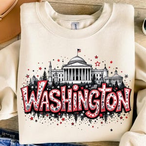 Pode incluir: Uma camisa de moletom de cor creme com uma estampa gráfica do horizonte de Washington D.C. O gráfico apresenta o Capitólio, o Monumento a Washington e outros marcos. A palavra "WASHINGTON" está escrita em letras vermelhas grandes com estrelas brancas e pequenas ilustrações em preto e branco de edifícios dentro das letras.