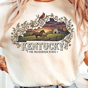 Op de afbeelding: Crèmekleurig T-shirt met een Kentucky-afbeelding. Het ontwerp bevat een zonsondergang, paarden en een schuur binnen de contouren van de staat. Een rode vogel zit op een tak boven de tekst "Kentucky, The Bluegrass State."