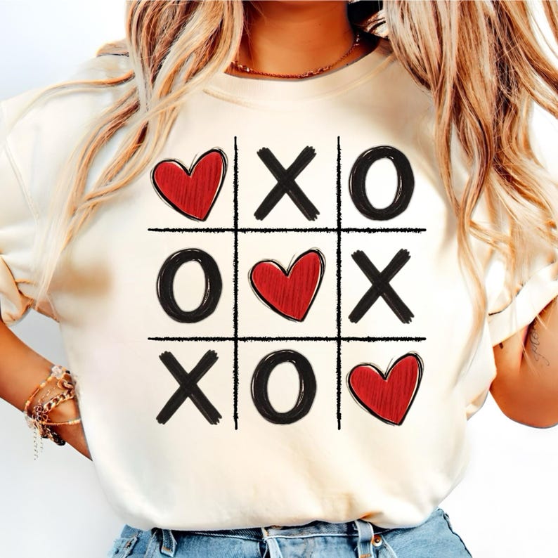 May include: White t-shirt with a black and red tic-tac-toe design featuring hearts instead of X's. The hearts are red and the tic-tac-toe board is black.