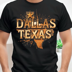 Puede incluir: Camiseta negra con las palabras "DALLAS TEXAS" en una fuente dorada desgastada. El diseño incluye una estrella dorada y un contorno del mapa de Texas, con un efecto de salpicaduras en el fondo. La camiseta es de cuello redondo clásico.