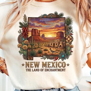 May include: Cream-colored t-shirt featuring a colorful graphic of a New Mexico landscape with a sunset, cacti, and a roadrunner. The text "NEW MEXICO THE LAND OF ENCHANTMENT" is printed below the image. The design is framed with decorative elements.