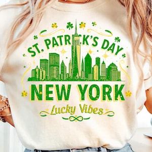 Puede incluir: Camiseta color crema con un diseño del Día de San Patricio. El diseño presenta un horizonte verde de la ciudad de Nueva York, el texto "St. Patrick's Day New York" y las palabras "Lucky Vibes". También se incluyen gráficos de tréboles y tréboles.