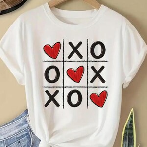 May include: White t-shirt with a black and red graphic of a tic-tac-toe board with hearts replacing the 'O's.