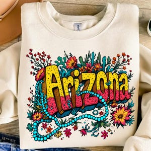 Puede incluir: Una sudadera beige con un diseño gráfico colorido de la palabra "Arizona" rodeada de flores y cuentas turquesa.