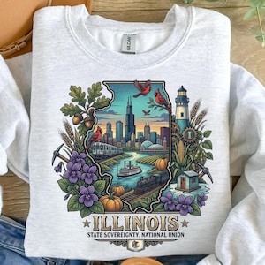 Op de afbeelding: Lichtgrijs sweatshirt met een kleurrijke Illinois staat grafiek. Het ontwerp bevat een stadsgezicht, trein, vuurtoren en de tekst "ILLINOIS STATE SOVEREIGNTY, NATIONAL UNION". De afbeelding is omlijst door bloemen, pompoenen en andere elementen.