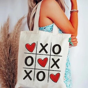 May include: A white canvas tote bag with a red heart and black X and O design. The design is a tic-tac-toe board with hearts replacing some of the X's and O's.