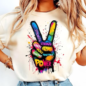 Puede incluir: Una camiseta blanca con un gráfico de signo de paz colorido. El signo de paz está formado por las palabras "Fuerza", "Unidad", "Paz", "Juntos", "Poder".