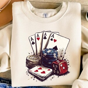 Puede incluir: Una sudadera blanca con un dise&ntilde;o gr&aacute;fico de cuatro cartas de juego, fichas de p&oacute;ker y un dado rojo. Las cartas son todas ases y tienen diferentes palos. Las fichas de p&oacute;ker est&aacute;n apiladas y tienen un dise&ntilde;o azul y blanco. El dado es rojo y tiene puntos blancos.