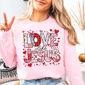 Könnte beinhalten: Rosafarbener Pullover mit Patchwork-Design, auf dem "Love like Jesus" steht, mit einer Schleife auf dem "O" von "Love" und umgeben von rosa Herzen.