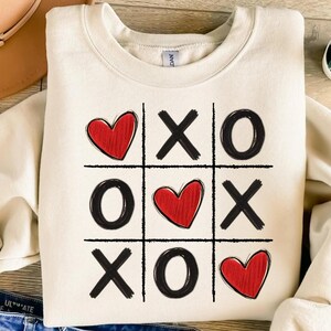 May include: A white sweatshirt with a black and red tic-tac-toe design. The design features hearts and X's and O's.