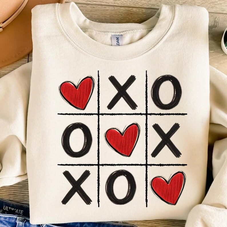May include: A white sweatshirt with a black and red tic-tac-toe design. The design features hearts and X's and O's.