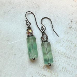 May include: A pair of handcrafted earrings featuring green rectangular beads and small, faceted, multi-colored beads. The earrings have dark metal ear wires and accents, with a vintage aesthetic. The beads are translucent, allowing light to pass through.