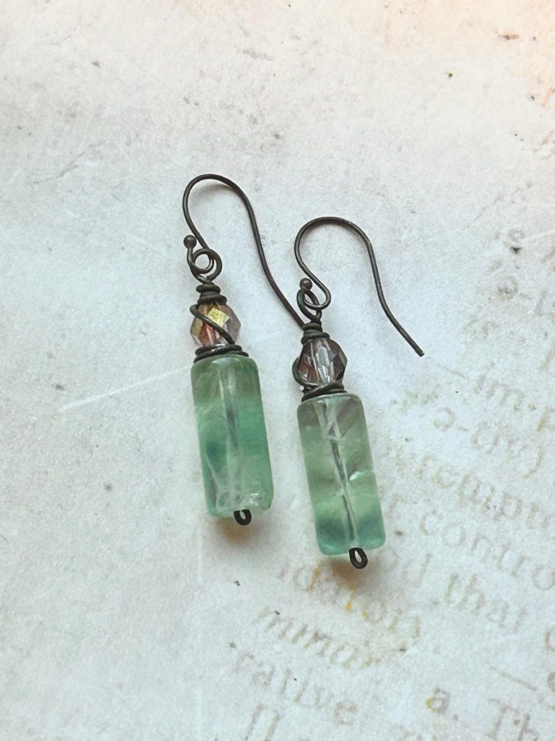 May include: A pair of handcrafted earrings featuring green rectangular beads and small, faceted, multi-colored beads. The earrings have dark metal ear wires and accents, with a vintage aesthetic. The beads are translucent, allowing light to pass through.