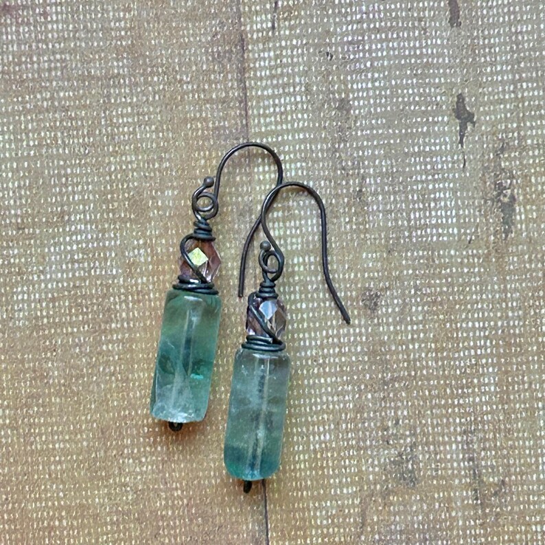 May include: A pair of handcrafted earrings featuring cylindrical, translucent green fluorite stones. Each earring has a faceted, light brown crystal accent and dark metal ear wires. The earrings are displayed on a textured, light brown surface.