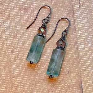 May include: A pair of handmade earrings featuring rectangular, translucent blue-green stones. The stones are accented with small, faceted brown beads and dark metal ear wires. The earrings are displayed on a textured, tan surface.