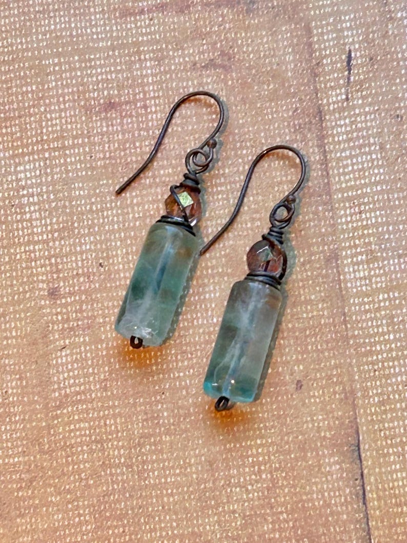 May include: A pair of handmade earrings featuring rectangular, translucent blue-green stones. The stones are accented with small, faceted brown beads and dark metal ear wires. The earrings are displayed on a textured, tan surface.