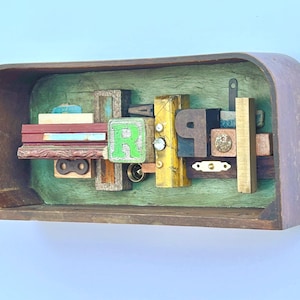 Puede incluir: Una caja de madera con un interior verde y un collage de madera, metal y pintura. El collage presenta las letras "R" y "P" y otros objetos diversos.