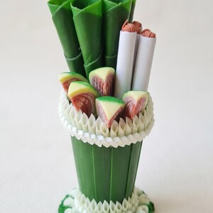 Pode incluir: Um arranjo decorativo em miniatura com folhas enroladas em verde escuro, dois bastões cilíndricos brancos e várias fatias triangulares pequenas com padrões rosa e verdes, numa base adornada com detalhes dobrados em branco e verde.