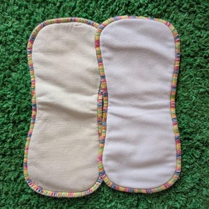 May include: Two rectangular cloth diaper inserts with a white interior and a colourful rainbow trim. The inserts are placed side by side on a green surface. The inserts are made of soft, absorbent material.
