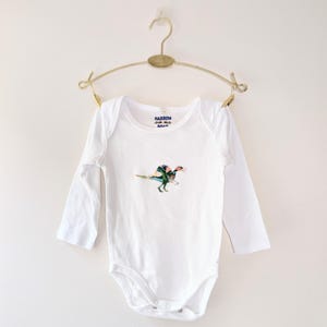 May include: White long-sleeved baby bodysuit featuring a colourful embroidered dinosaur. The bodysuit is suspended from a gold-toned hanger. The text "HARROM" is printed on the neckline.