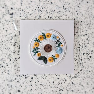 Op de afbeelding: Ronde geborduurde patch met een witte madelief met een bruin hart, omringd door gele en blauwe bloemen en groene bladeren. De patch is op een witte kaart.