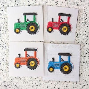 Puede incluir: Cuatro parches bordados de tractores en verde, rojo, naranja y azul. Cada tractor tiene un techo negro, una cabina blanca y un neumático negro con centro amarillo. Los parches están sobre tarjetas blancas.