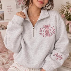 May include: A light gray quarter-zip sweatshirt featuring pink embroidery on the chest depicting ballet slippers tied with a bow, surrounded by floral sprigs and small bows. The left sleeve is embroidered with the name Hailey and a small heart.