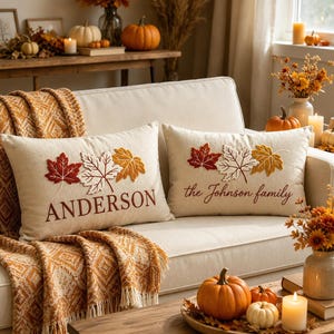 Puede incluir: Dos cojines rectangulares de color beige con hojas de otoño bordadas en rojo, blanco y dorado. Uno muestra el texto ANDERSON, mientras que el otro presenta el texto the Johnson family en letra cursiva, ambos centrados bajo las hojas.