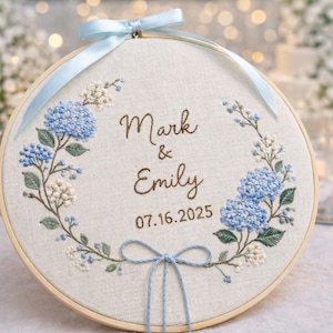 Könnte beinhalten: Gestickter Stickrahmen mit den Namen "Mark & Emily" und dem Datum "07.16.2025". Das Design umfasst blaue Hortensien und weiße Blumenstickereien, mit einer hellblauen Schleife. Der Rahmen befindet sich vor einem verschwommenen Hintergrund.