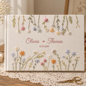 Pode incluir: Um álbum de fotos retangular coberto com tecido branco, apresentando um bordado floral intrincado em tons pastel de rosa, amarelo, azul e verde. A capa exibe os nomes bordados Olivia e Thomas com a data 18.05.2025 em fio rosa.