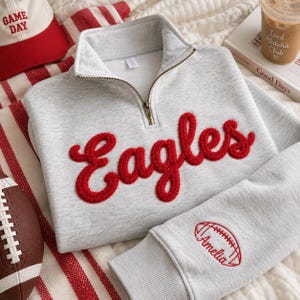 May include: A light gray quarter-zip sweatshirt featuring the word Eagles in red textured chenille lettering across the chest. The sleeve includes an embroidered red football graphic with the name Amelia written in cursive script underneath.