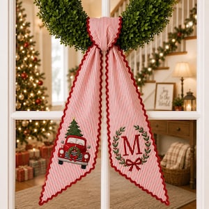 May include: A red and white striped fabric door hanger featuring a green boxwood wreath at the top. The left side displays an embroidered red truck carrying a Christmas tree, while the right side shows a green wreath with a red letter M.