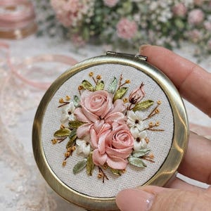 Könnte beinhalten: Ein runder Kompaktspiegel im antiken Stil mit floraler Stickerei. Die Stickerei zeigt rosa Rosen aus Band, weiße Blüten und grüne Blätter auf cremefarbenem Stoff. Der Spiegel hat einen goldfarbenen Metallrahmen.