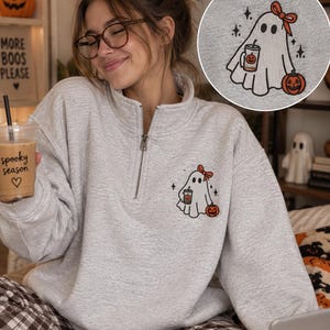 May include: A light gray quarter-zip sweatshirt featuring an embroidered design of a ghost wearing a red bow, holding a coffee cup with a pumpkin print, and standing next to a jack-o'-lantern. The embroidery includes small black star accents.