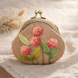 Puede incluir: Un monedero de tela color beige con un cierre metálico, con flores rosas bordadas y hojas verdes. El monedero tiene forma redondeada y una estética vintage, perfecto para llevar objetos pequeños.