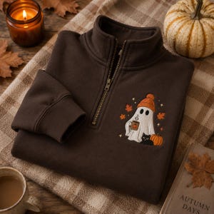 Peut inclure: Un sweat-shirt marron foncé à fermeture éclair quart, orné d'une broderie représentant un fantôme blanc avec un bonnet orange, tenant une tasse à motif citrouille, accompagné d'un chat noir, d'une citrouille et de feuilles d'automne.