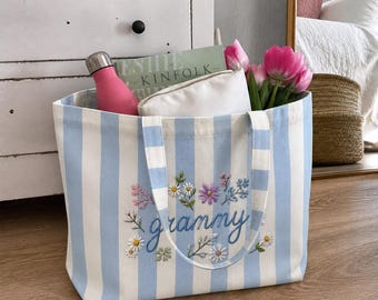 Pode incluir: Uma bolsa de lona com listras azul-claras e brancas, com detalhes florais bordados e a palavra "grammy" em letra cursiva azul. A bolsa contém uma garrafa de água rosa, uma bolsa branca e um buquê de tulipas rosas.