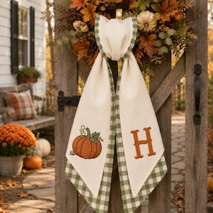 May include: A cream-colored fabric door hanger featuring a green and white gingham border. One side displays an embroidered orange pumpkin with green leaves, while the other side features a large, embroidered orange letter H in a serif font.