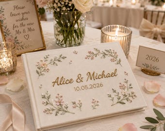 Peut inclure: Un livre d'or en tissu crème orné de brins de fleurs brodés dans des tons roses et verts. La couverture présente le texte Alice & Michael brodé en doré ainsi que la date 10.05.2026. Un stylo doré est posé horizontalement devant le livre.