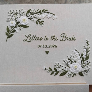 May include: A rectangular off-white fabric-covered book featuring embroidered white floral arrangements in the top-left and bottom-right corners. The text "Letters to the Bride" is stitched in green, followed by the date "07.10.2026" and a small heart.
