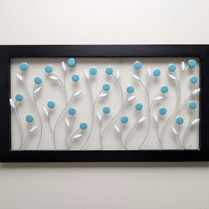 May include: A rectangular black frame containing a wall art piece with thin silver wire stems, small white leaves, and round light blue circular accents arranged in a floral pattern against a light cream background.