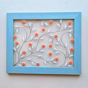 May include: A rectangular wall art piece with a light blue wooden frame. Inside, a design of black, curved lines with white leaves and orange circular accents creates a floral pattern. The background is a light beige color.